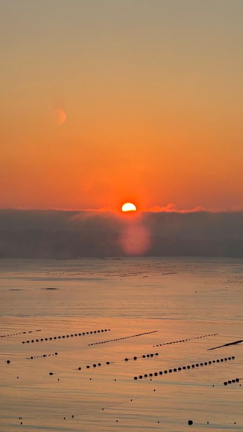 Sunset over a calm sea with an orange sky; the sun is just above the horizon, reflecting on the water, with lines of buoys on the surface.