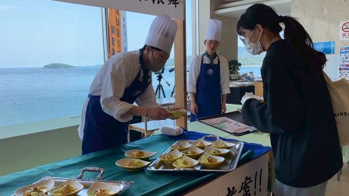 Chef in a tall white hat and blue apron serves bowls of food to a customer at an outdoor seaside counter, with large windows and ocean view in the background.