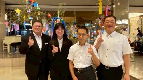 Four colleagues in business attire pose in a decorated lobby, giving thumbs up with smiles. The festive background hints at a celebration or event.