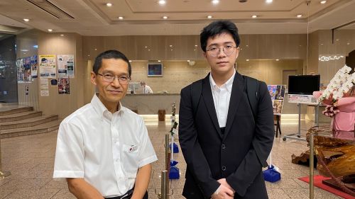 Two men pose in a lobby: an older man in a white shirt on the left and a younger man in a black suit on the right, with posters and a reception desk behind them.