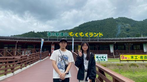 Two people pose on a wooden bridge in front of a building with a sign reading '道の駅 もくもくランド' and a hillside in the background.