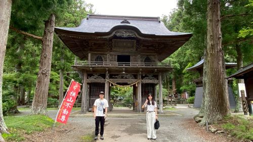 Two people stand in front of a large wooden gate to a Japanese shrine, surrounded by tall trees and a stone path.