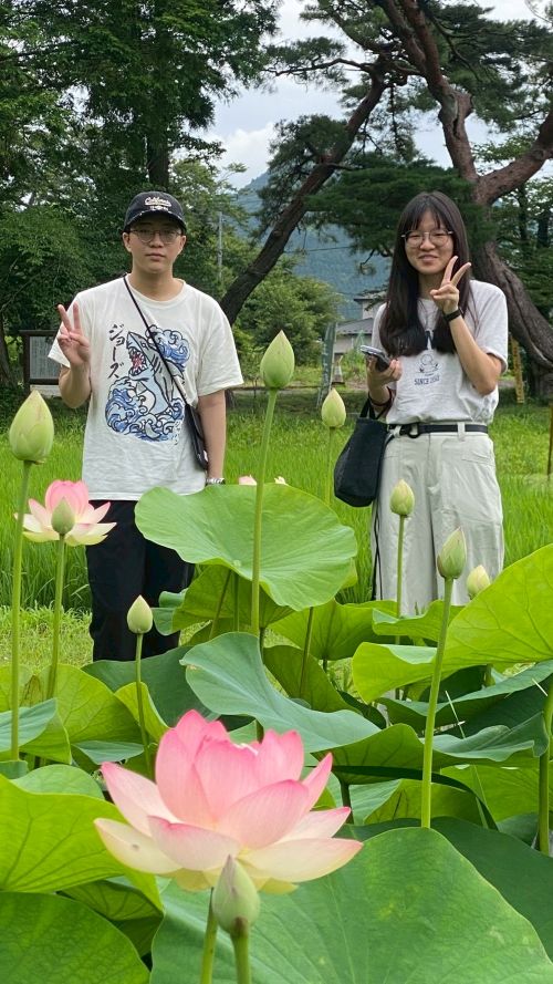 Two people stand among blooming lotus plants in a grassy garden, posing with peace signs while pink lotus flowers fill the foreground.