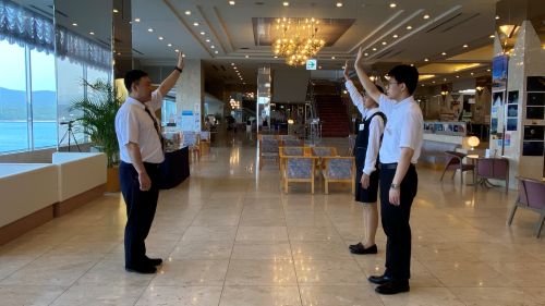 Three hotel staff in uniform stand in a bright lobby facing each other and saluting with raised right arms, chandeliers overhead.