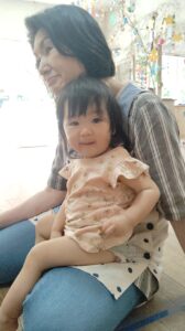 Woman sitting with a smiling toddler on her lap indoors, colorful hanging crafts in the background.