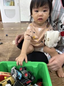 Toddler sitting on someone's lap, playing with toy cars in a green plastic bin on a wooden floor.