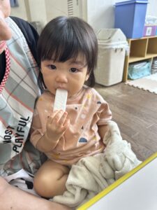 Young child sitting on an adult's lap, chewing a white popsicle, wearing a peach shirt with colorful shapes.