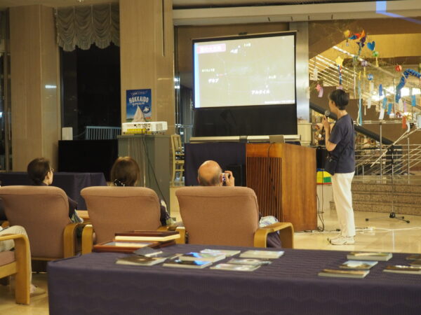 Speaker with microphone presenting to an audience in front of a projected display in an indoor space.