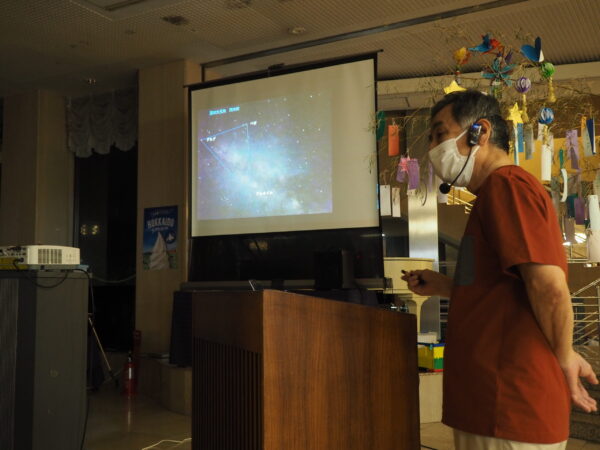 Presenter wearing a mask and headset stands at a lectern, projected star-chart visible on a screen behind him in a dim room.
