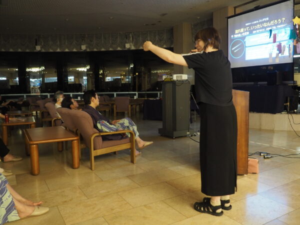 Presenter in a black outfit on stage, arms extended as she speaks to an audience in a conference room with slides on a large screen to the right.