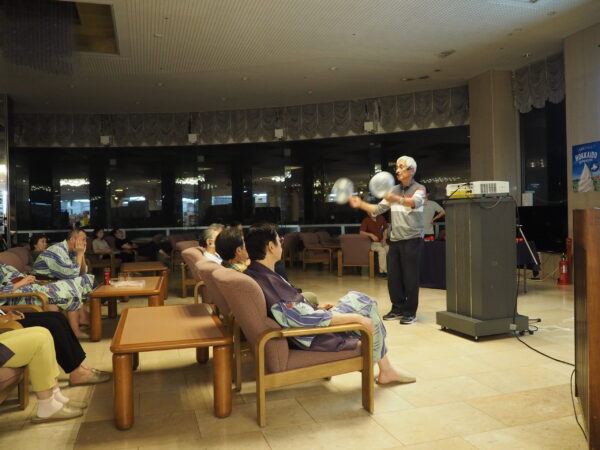Older man in a striped shirt juggling two balls in a seated audience area, projector on a podium nearby, a small crowd watching.