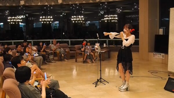 Young female singer on stage with microphone, performing for an audience in a lounge-style venue.