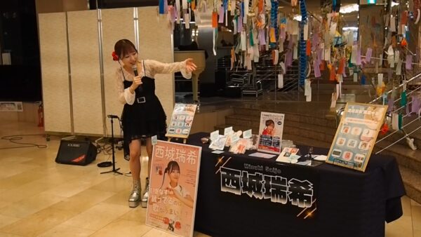 Female singer performs with a microphone at a mall kiosk, promotional posters and a decorated booth nearby.
