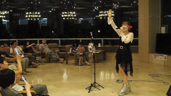 Young female singer with microphone performing on stage, one arm raised, audience seated and watching in a lit lobby area with glow sticks