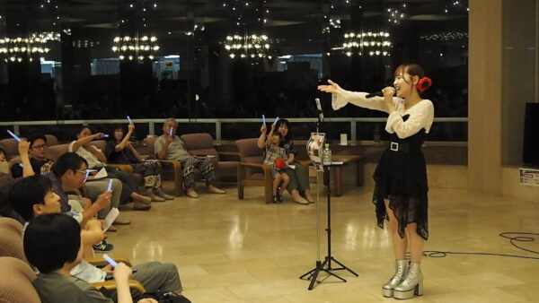 Female singer on stage holding a microphone, performing to an audience waving blue light sticks in a dim, indoor venue.