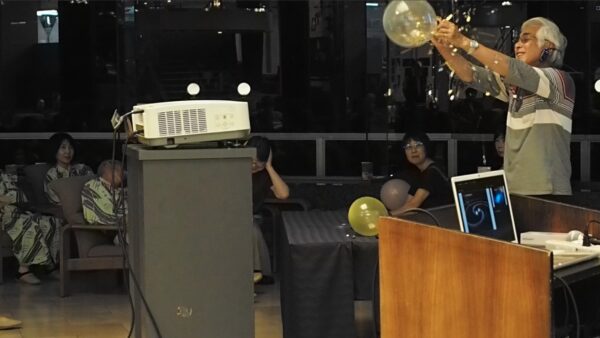 Older man with headphones and a striped shirt releases a balloon during a party as attendees watch nearby.