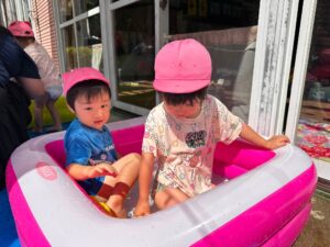 Two children wearing pink hats sit in a pink and white inflatable pool outdoors beside a glass storefront nearby.