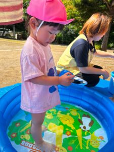 Young child wearing a pink cap and light pink shirt standing beside a blue inflatable kiddie pool outdoors.