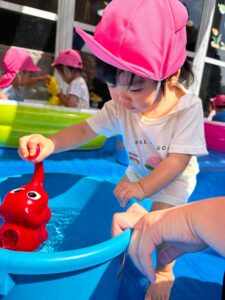 Child wearing a pink hat plays with a red toy in a blue water tub, with other kids in pink hats in the background.