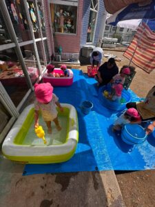 Children wearing pink hats playing with water in small plastic pools on a blue tarp, supervised by adults outside a colorful building window decorations are visible in the background.