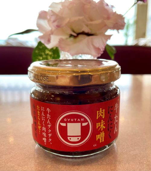 Jar of Japanese gyutan beef tongue miso paste with a metal lid, orange-red label, on a wooden table; blurred pink flowers in the background.