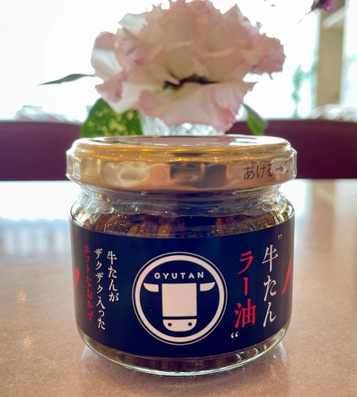 Glass jar of Gyutan sauce with a blue label and gold lid on a kitchen counter, pink flower in the background.