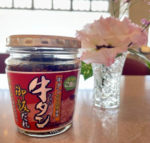 Glass jar of Japanese gyūtān tare (beef tongue sauce) with a copper lid on a table, beside a crystal vase of pink flowers.