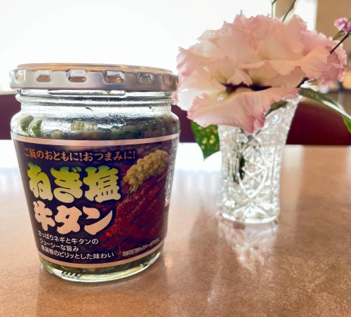 Jar of Japanese negi shio tare sauce on a table, with a crystal vase of pink carnations in the background.
