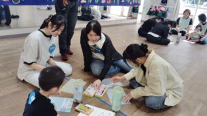 Group of kids and adults sitting on a wooden floor, coloring and drawing with crayons and paper, surrounded by craft supplies in a bright, classroom-like space.
