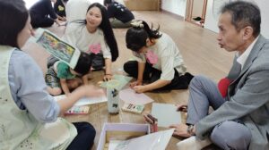 Group of kids and adults on a wooden floor doing crafts with paper and crayons, supervised by a man in a gray blazer.