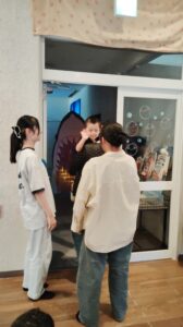 Child waves at camera while two adults stand near a glass door with a large pink shark cutout inside behind them growling in a doorway.