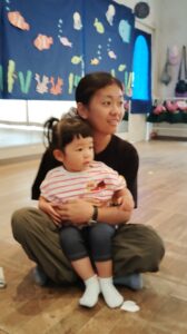 Mother and toddler daughter sit on the floor together, the child resting in her lap, both looking to the right in a room with blue sea-themed decorations behind them.