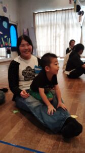 Two kids sit on a wooden classroom floor, a smiling girl with a blue name tag behind a younger boy in a black shirt.