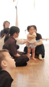 Toddler stands with help from adults during a group activity in a bright room with others on the floor.