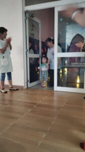 Young child in plaid shorts and red shoes holds hands with an adult while stepping through a glass door beside a clapping person in a hallway.