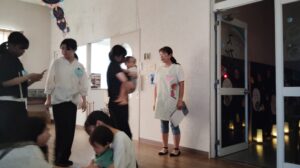 Group of adults and children in a hallway; a caregiver holds a baby while others chat nearby.