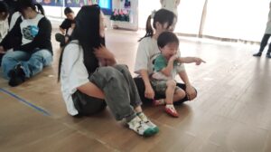 Group of kids and adults sitting on a wooden gym floor, with a smiling child pointing and a laughing toddler on a caregiver's lap.