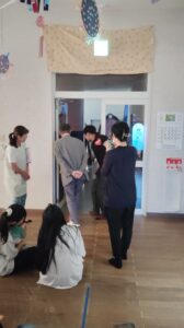 Adults stand by an open classroom door while children sit on the wooden floor, watching something inside.] ,
