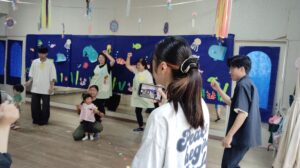 Adults and children pose and dance in front of a blue ocean-themed backdrop with hanging decorations; a toddler sits with a caregiver on the floor while a person records with a smartphone in the foreground.