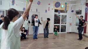 Group of adults and children participating in a classroom activity with arms raised, a dancing toddler in green shorts, and colorful decorations overhead.