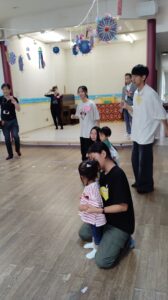 People gather in a decorated indoor hall; a woman kneels beside a young girl while other adults and children stand or sit nearby, participating in a small event or activity. Colorful hanging crafts dangle from the ceiling as a piano and tables are in the background.