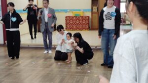 Adults and a child participate in a group activity in a community hall, with colorful paper shapes scattered on the floor.