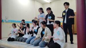Group of students on a stage, kneeling and standing, wearing blue heart name tags, performing with hand gestures.