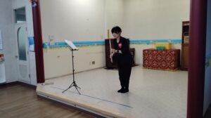 Woman in black performs a flute piece on a small stage, with a music stand and sheet music nearby in a decorated room.