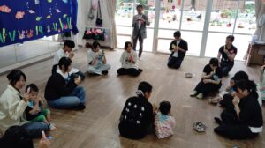 Group of adults and children sitting in a circle on a wooden floor, eating colorful popsicles at an indoor event, with a blue sea-themed backdrop and large windows decorated with cartoon decals.
