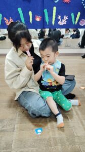 Woman and child sitting on a wooden floor, both eating pink popsicles, with a blue underwater-themed backdrop behind them.