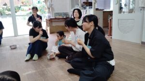 Group of adults and a young child sitting on a wooden floor, eating snacks from small cartons in a bright room with window decorations
