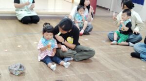 Group of adults and young children sitting on a wooden floor, feeding themselves snacks in a circle.