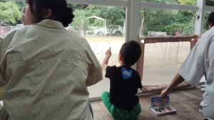 Child wearing a blue heart sticker reading ゆず sits and draws on the window of a bus or tram, with two adults nearby and a park outside.