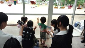 Children and adults drawing on a large glass window decorated with sea-themed stickers, while looking outside at a sunny yard. A group of kids with markers sketch on the window as caregivers supervise.